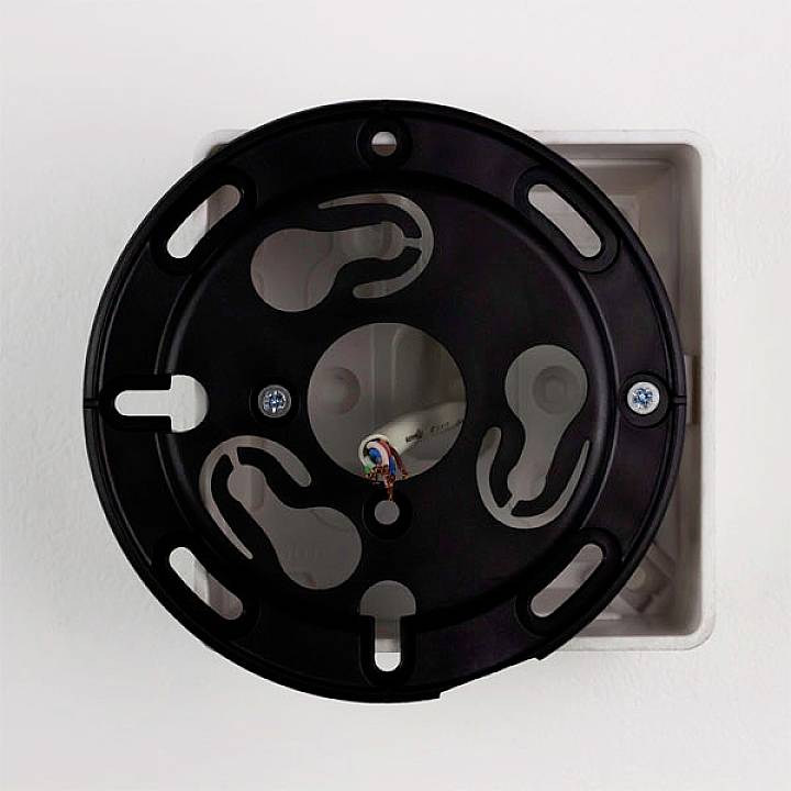 A round, black mounting plate on a white wall, with several openings and cables visible in the centre.