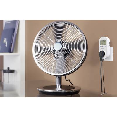 A desk fan is standing on a table in front of a brown wall. To the right, a digital control unit is mounted on the wall.