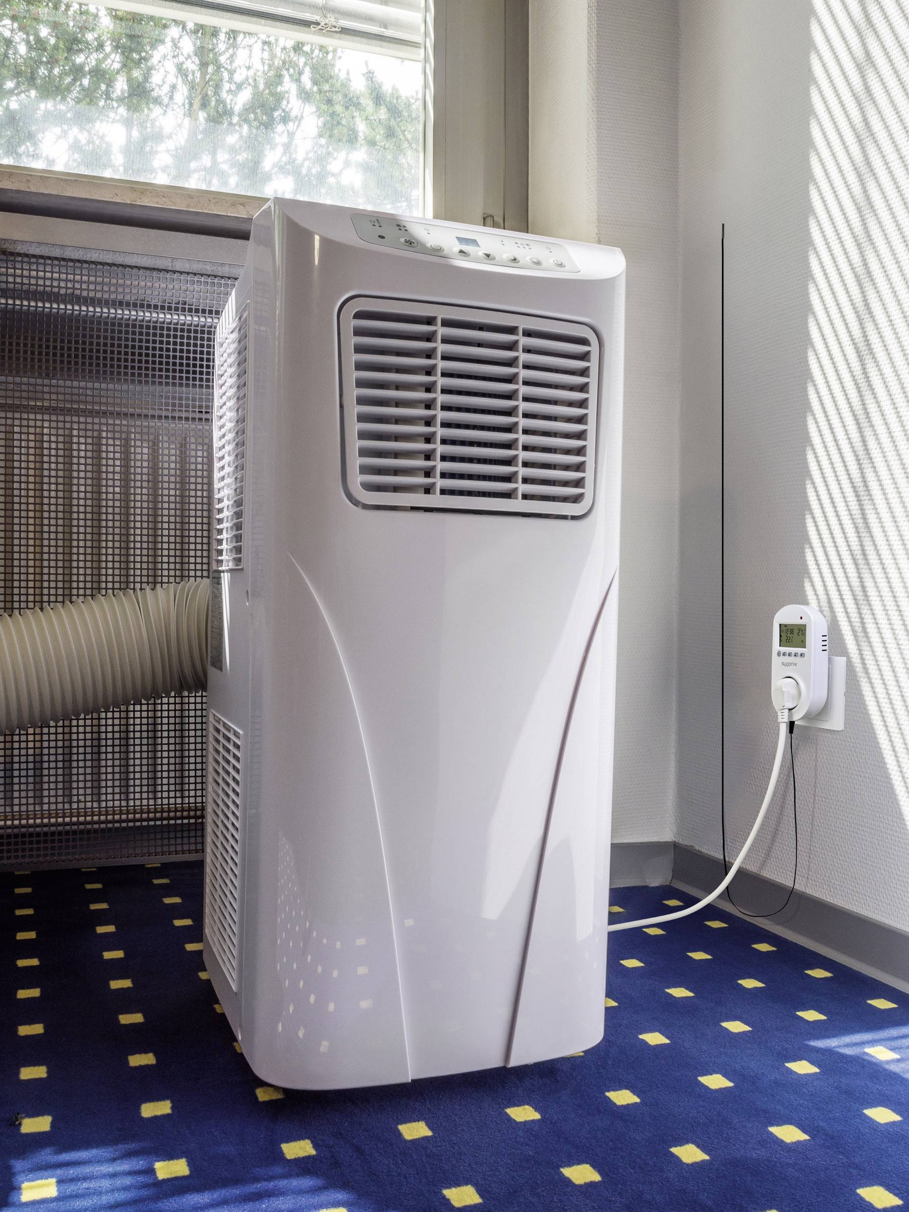 A portable air conditioning unit stands in a room with blue carpet. A hose is connected to the device and leads to a window.