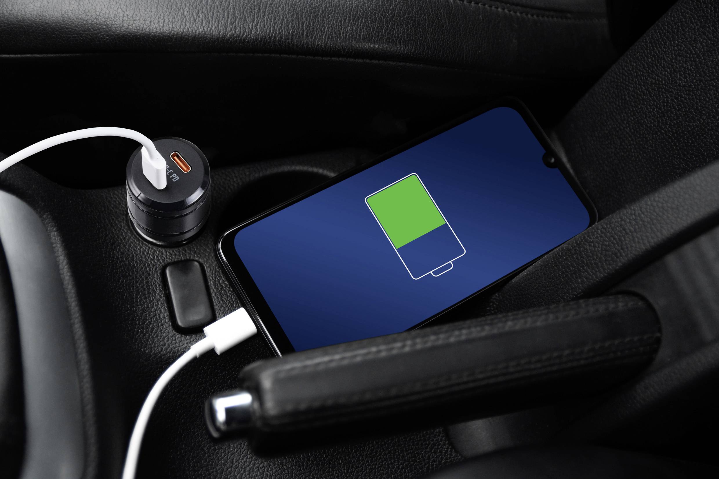A smartphone is lying on a car seat displaying a green charging bar. It is connected to a charger plugged into the cigarette lighter.