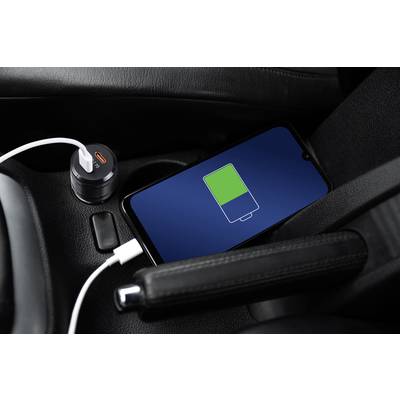 A smartphone is lying on a car seat displaying a green charging bar. It is connected to a charger plugged into the cigarette lighter.