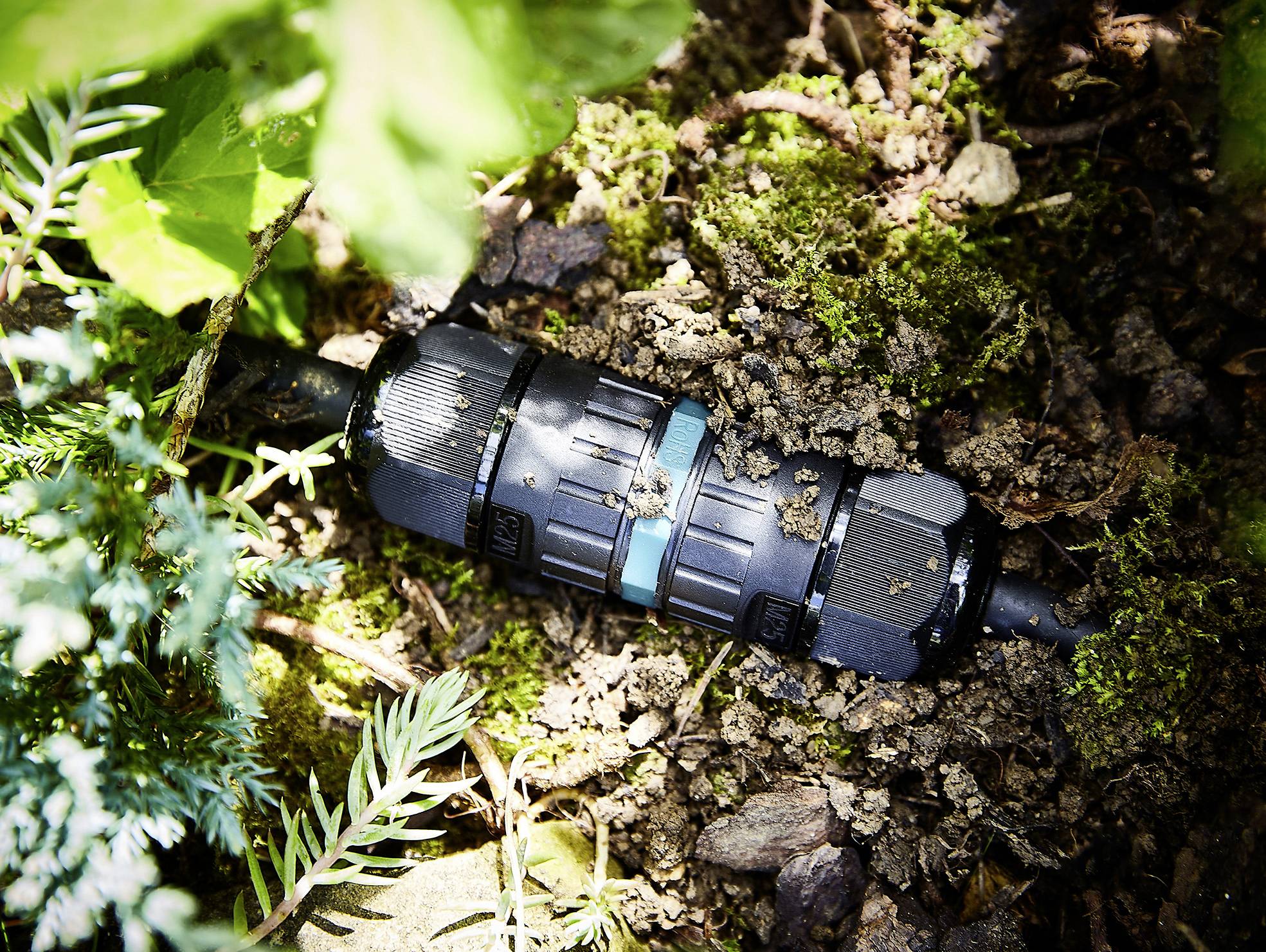A black cable connector lies on an earthy ground surrounded by plants. It connects two cables to each other.