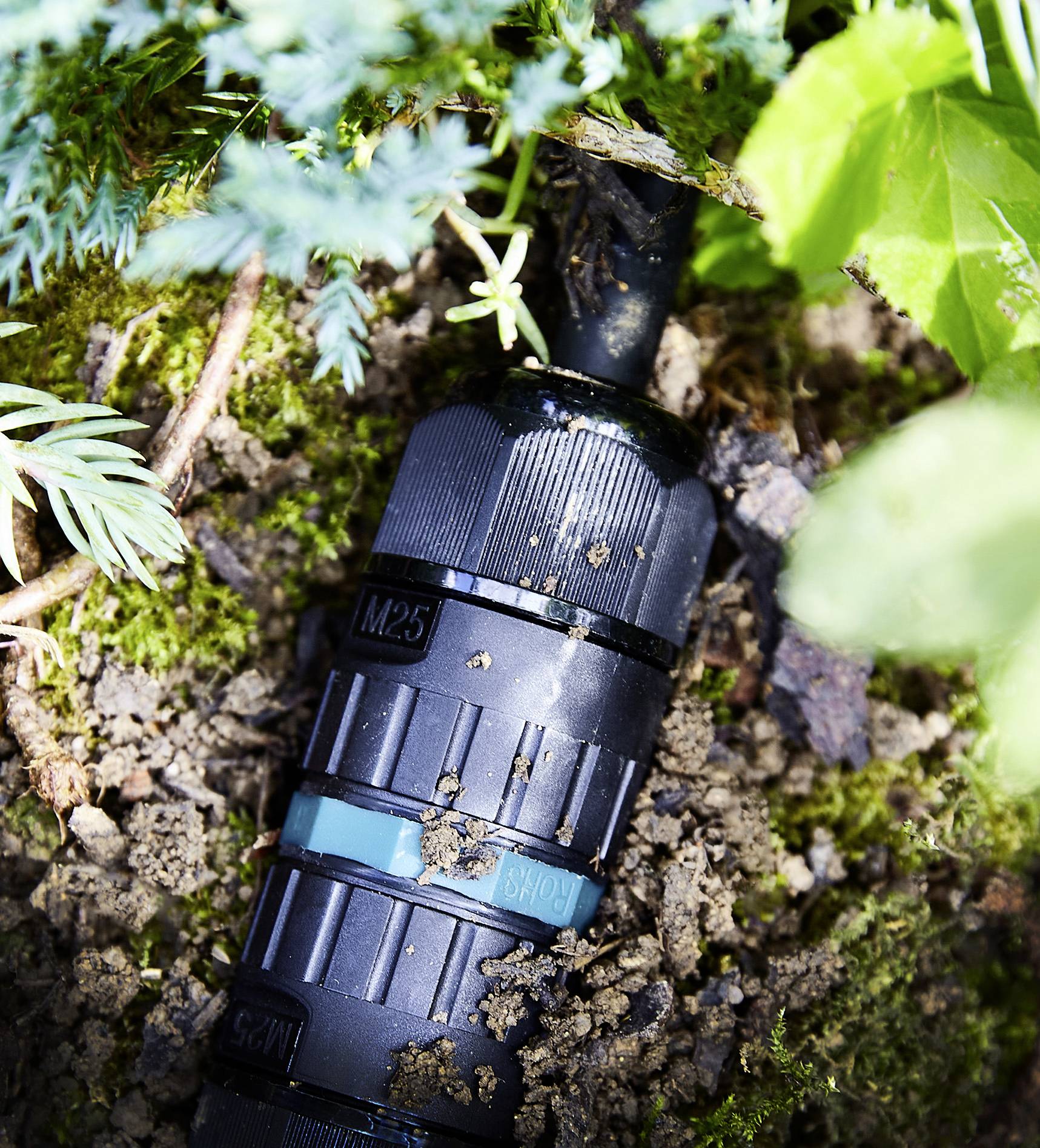 A black water pipe running through earth and green plants, visibly connected with a label reading 'M25'.