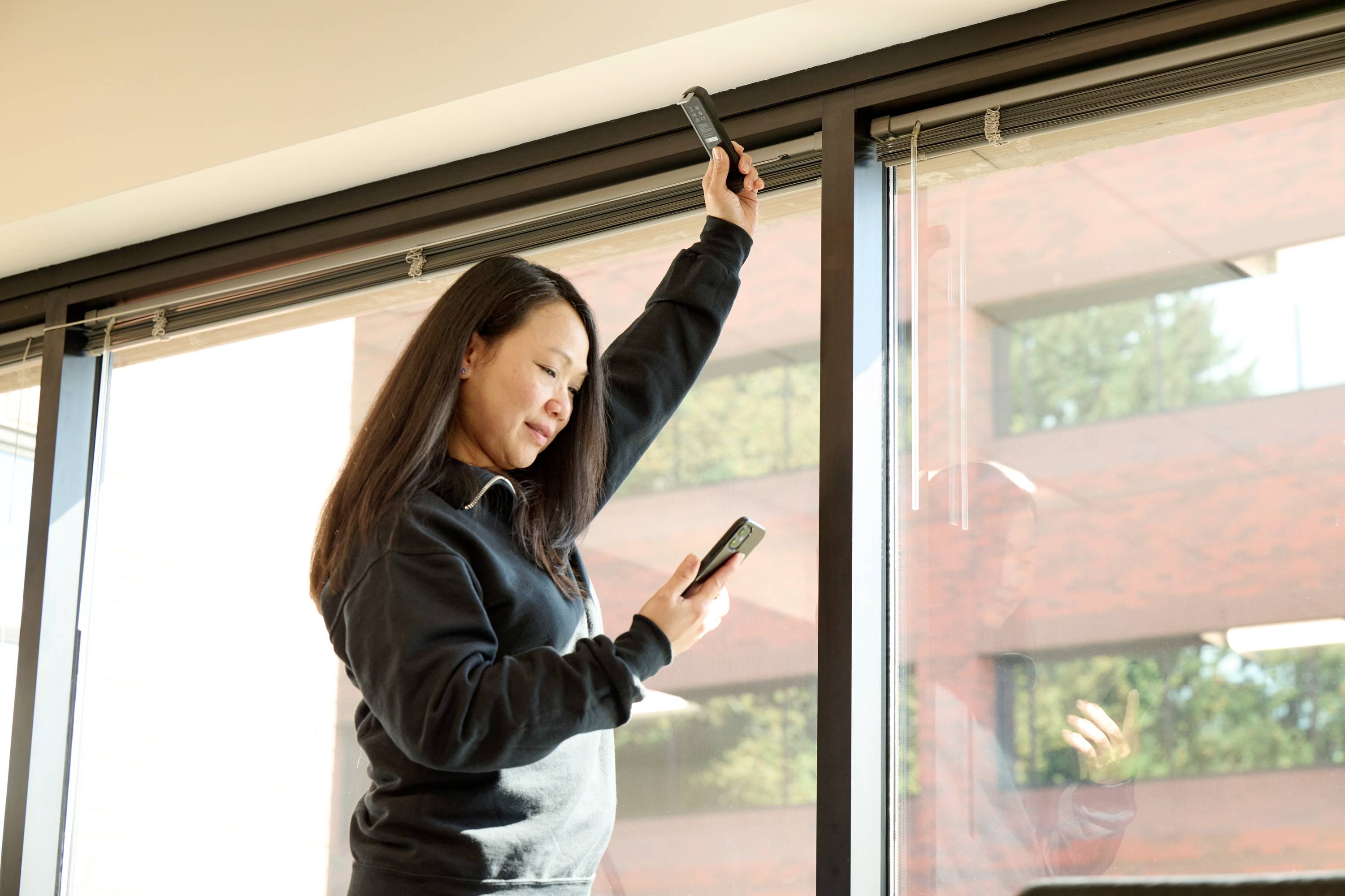 A person is standing in a room using a smartphone and a remote control to operate the blinds of a large window.