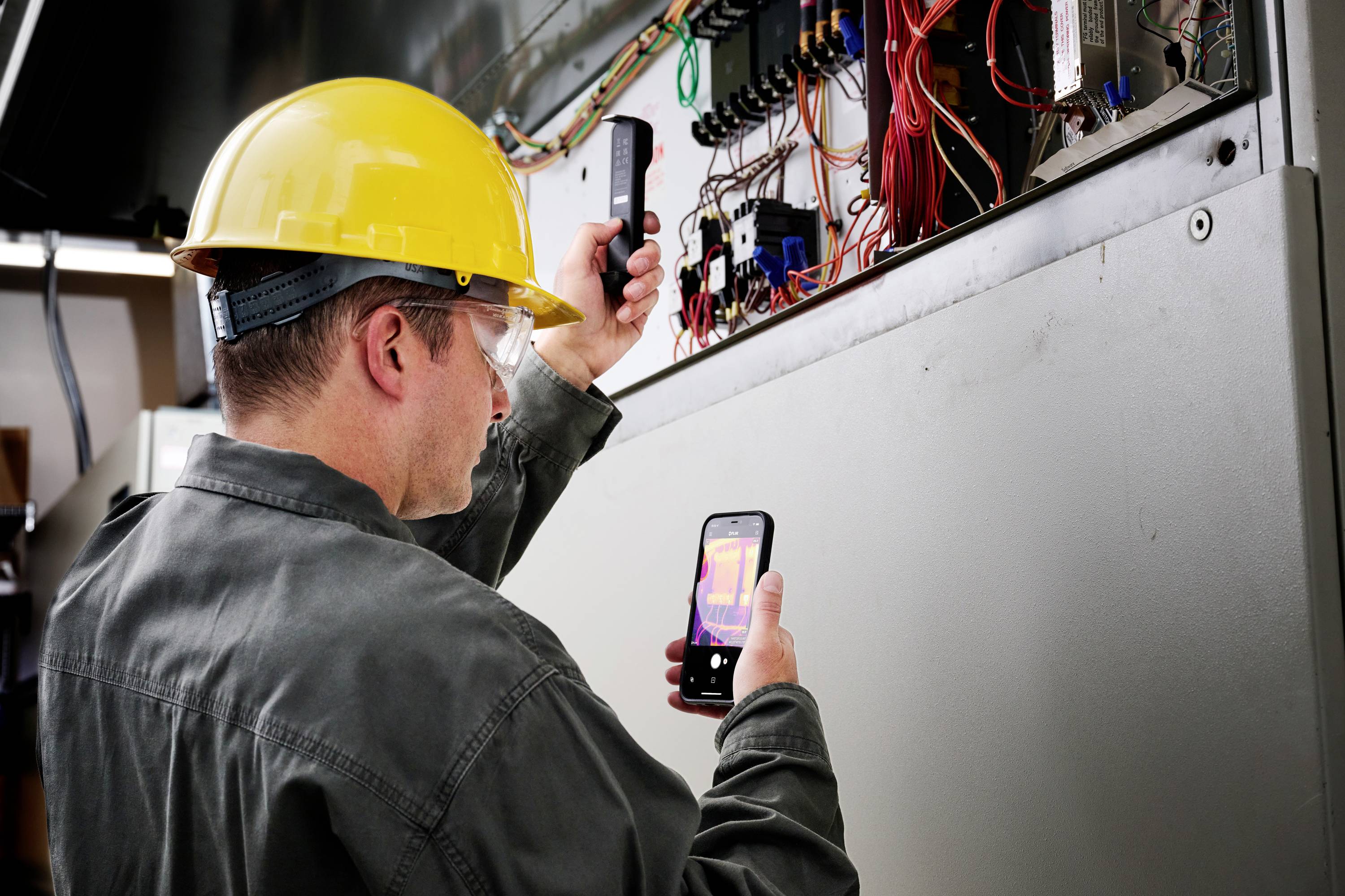A technician wearing a safety helmet and glasses is examining a control panel with a thermal imaging camera to analyse temperature variations.