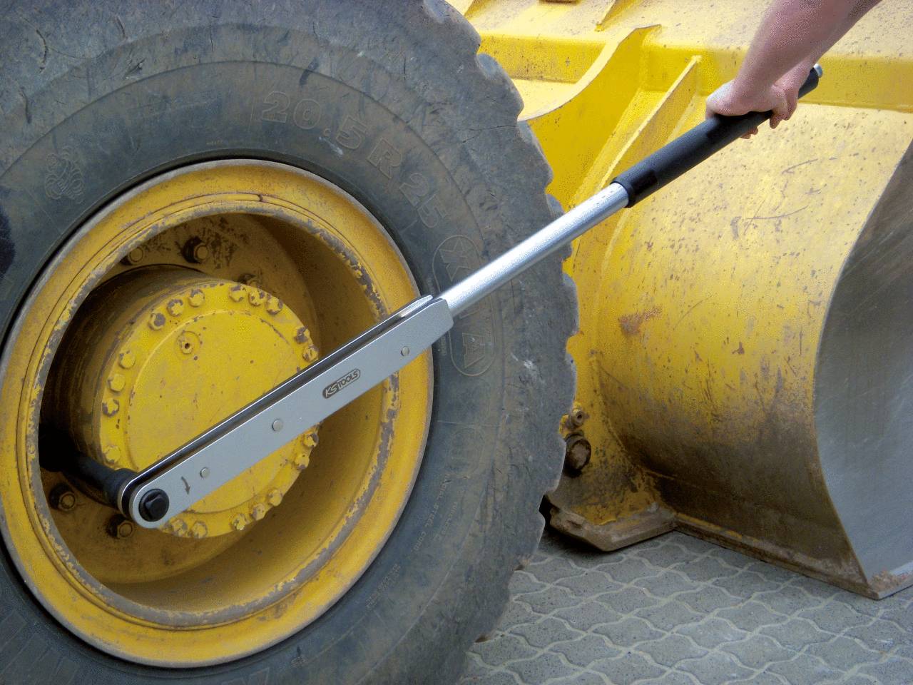 A man is using a long torque wrench to loosen the wheel nut of a large yellow construction vehicle on a paved surface.