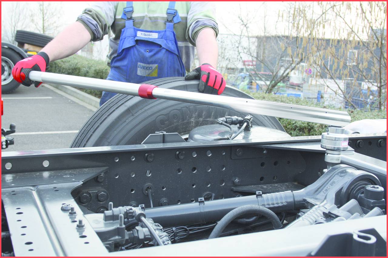 A person wearing blue workwear is using a long pole to inspect the tyres on a lorry chassis. Trees can be seen in the background.