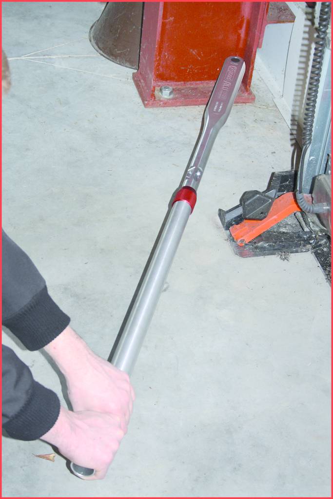 A person is using a large torque wrench to work on a vehicle. The floor is concrete, with workshop equipment in the background.