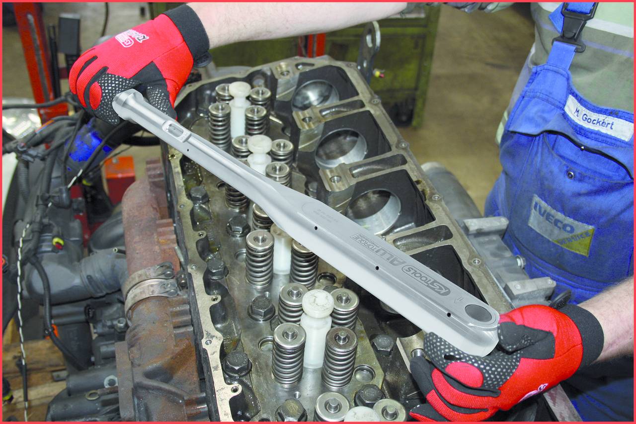 A person wearing red gloves is working with a torque wrench on an engine block, tightening cylinder head bolts.