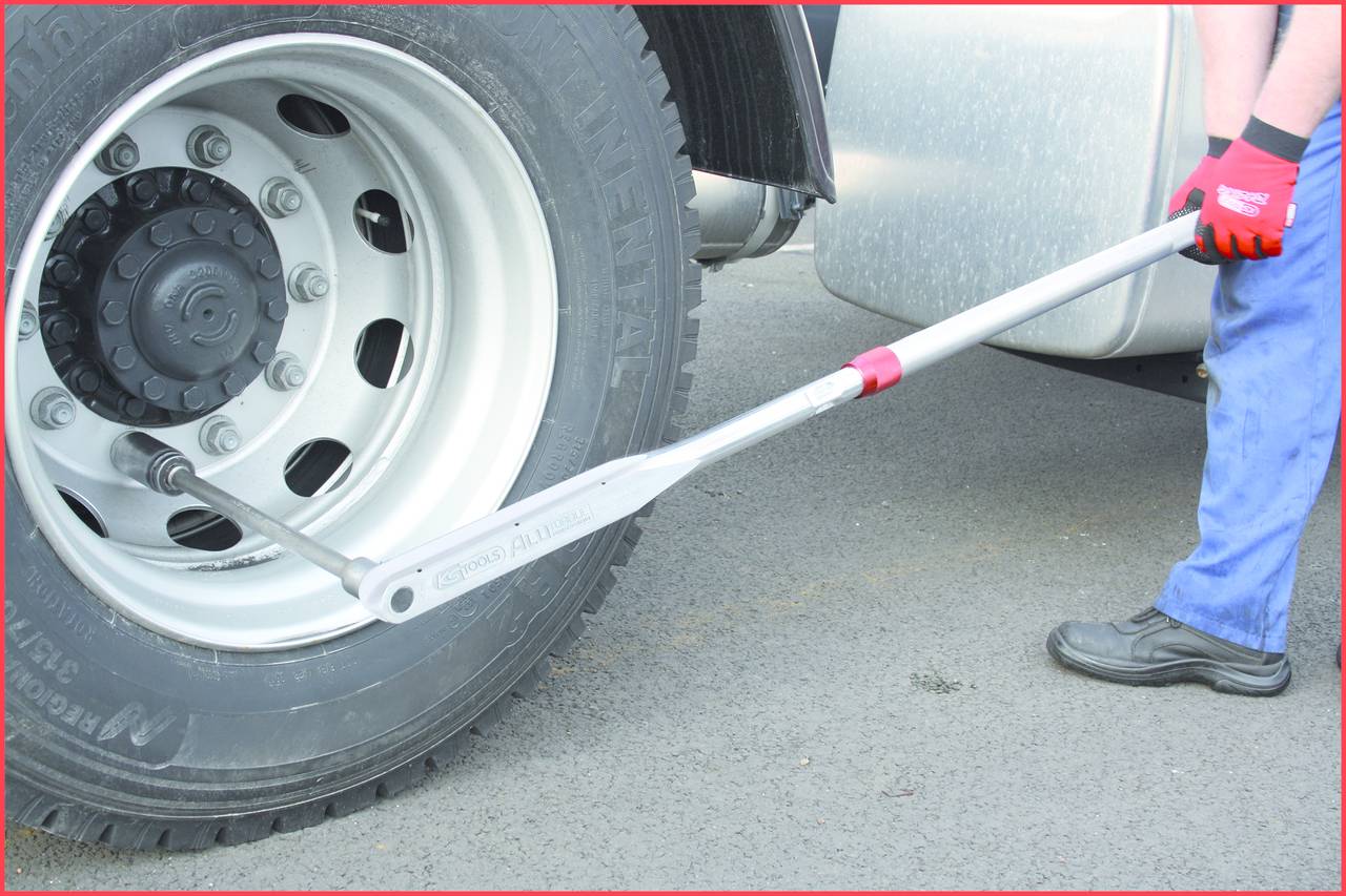 A person is using a torque wrench to tighten a lorry wheel. The tool is positioned on a wheel nut, with a road in the background.