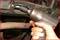 A hand holds an electric ratchet and tightens a screw on a car. The surroundings are dirty and industrial.