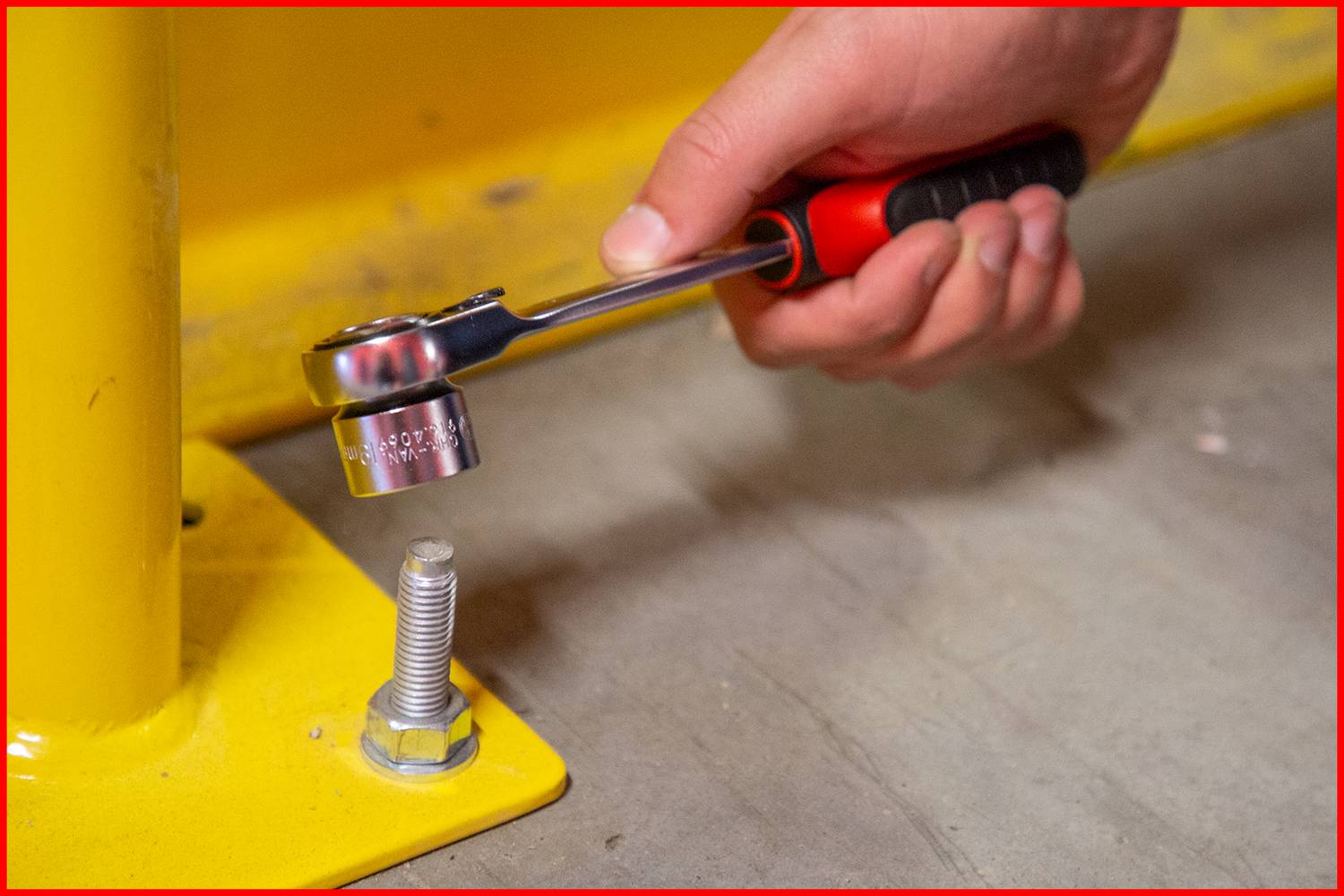 A hand is tightening a screw through a yellow metal plate using a ratchet.