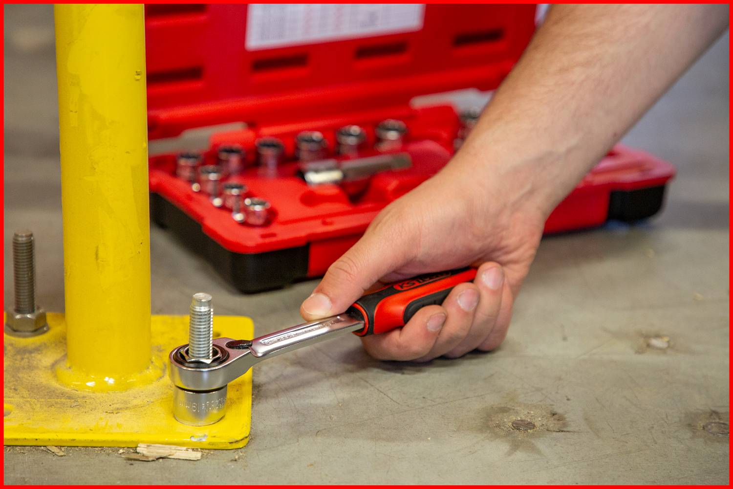 A person is tightening a screw on a yellow post with an adjustable spanner. In the background, a red toolbox lies nearby.