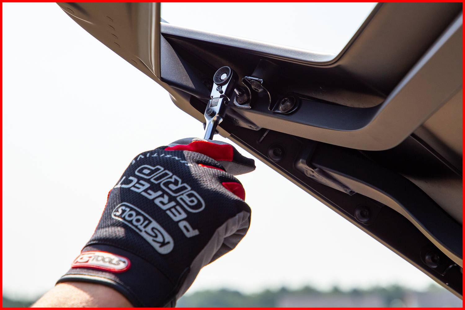 A hand in a work glove assembles a car part with a ratchet, with sunlight in the background.