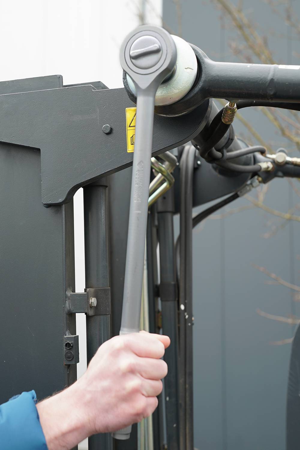 A hand is gripping a large spanner, which is positioned against a substantial metal bolt. The background displays machine components.