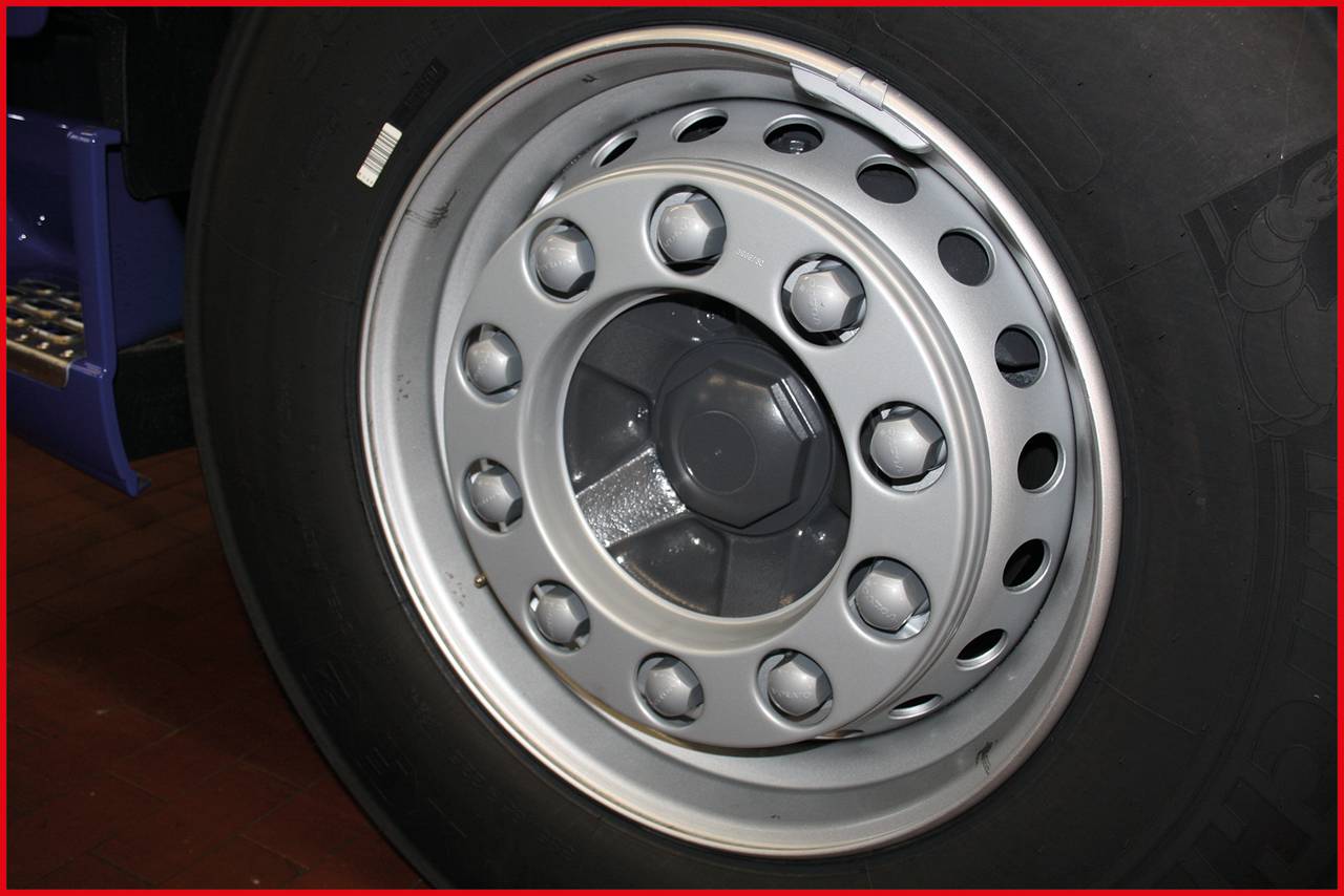 Close-up of a lorry tyre with a silver wheel rim featuring multiple nuts. The tyre is standing on a dark red surface.