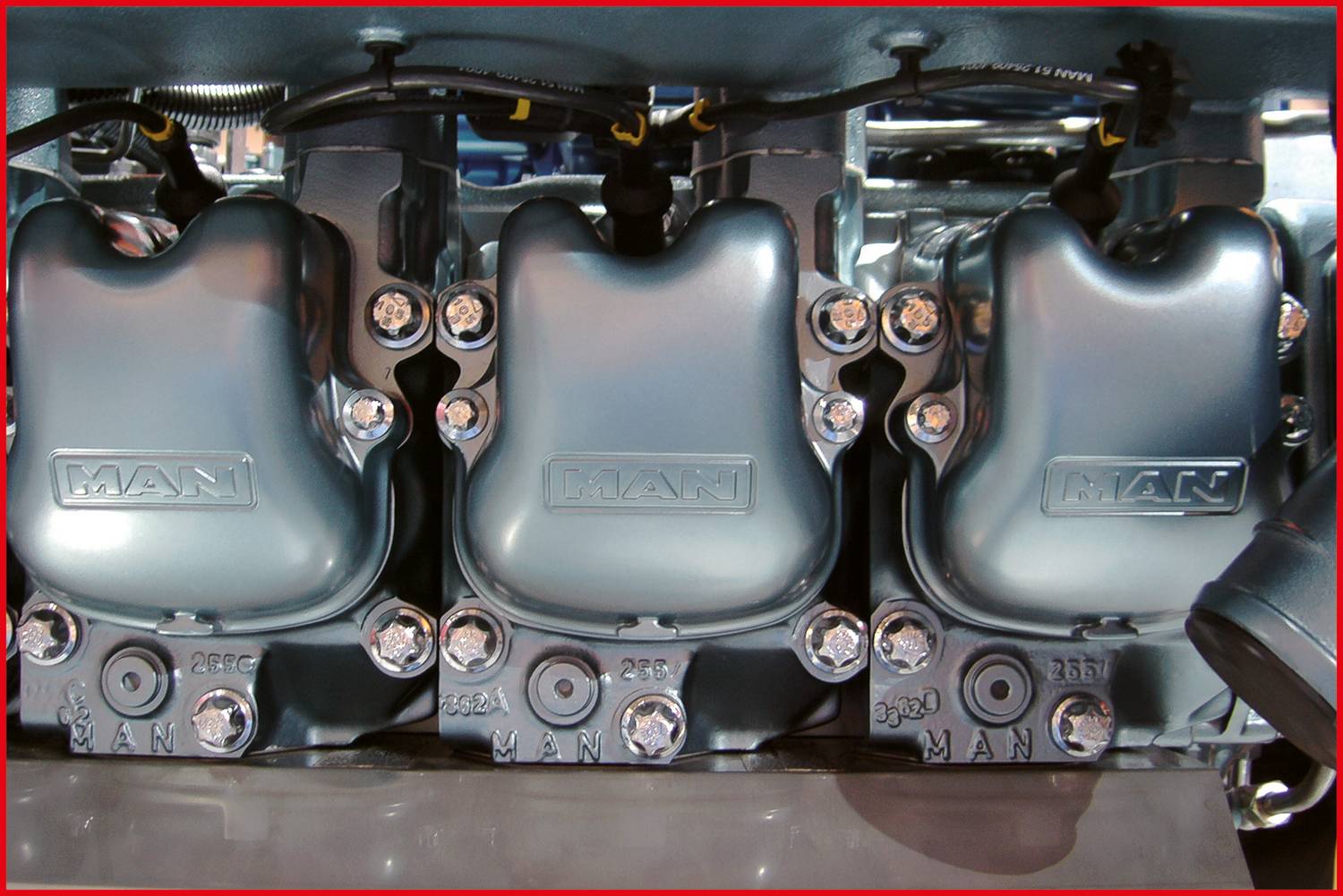 A MAN engine block with three silver cylinder covers lying side by side, with visible bolting and wiring.