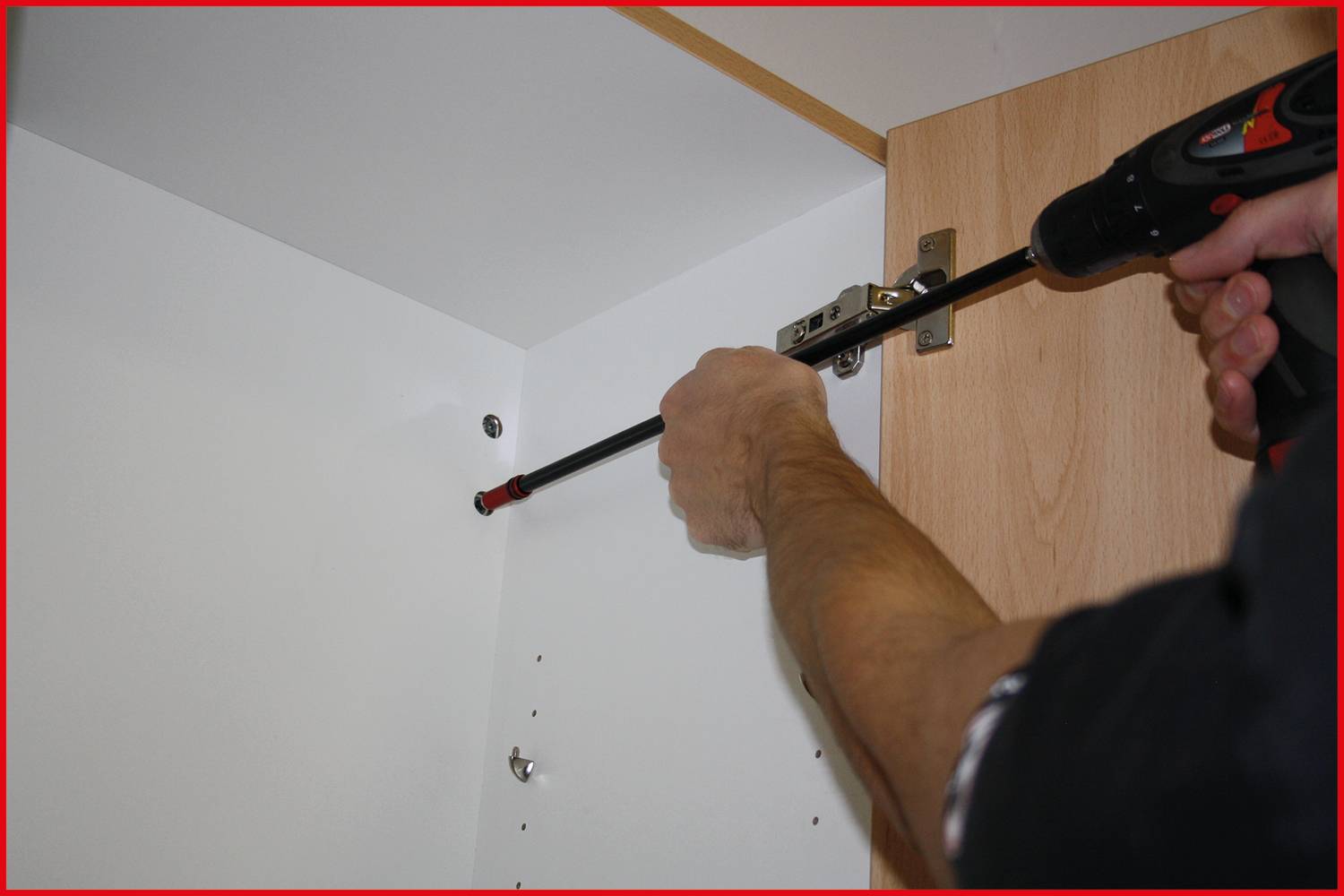 A person is attaching a hinge to a wooden cabinet with a black cordless drill in a white interior.