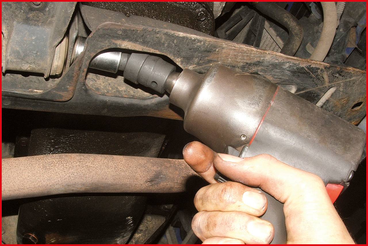 A person is holding a pneumatic impact driver and loosening a screw on a vehicle undercarriage. The surroundings show vehicle components.