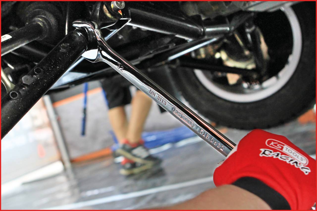 A person wearing red gloves is repairing a vehicle, using a spanner to work on the chassis. Tyres are visible in the background.