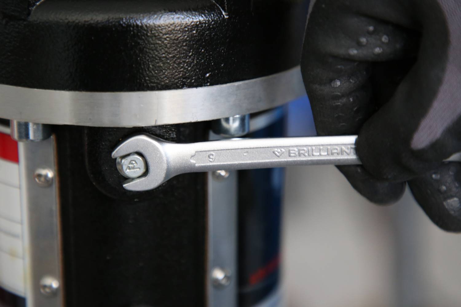 A hand wearing a glove is holding a spanner to tighten a screw on a metal machine.