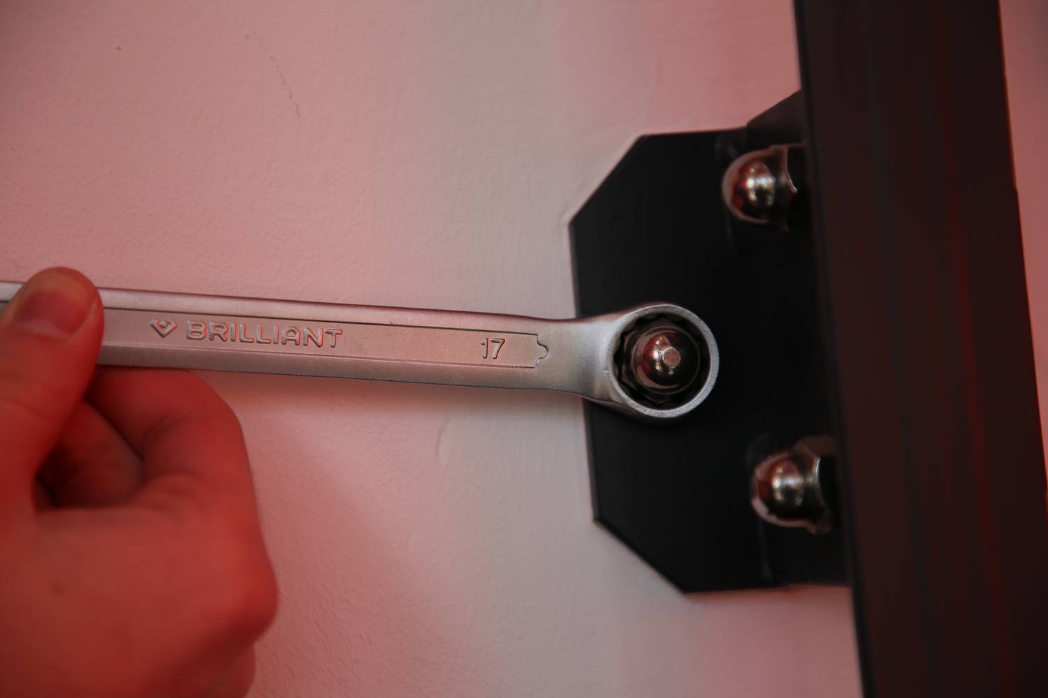 A person is holding a spanner to tighten a screw on a black bracket. The spanner indicates a size of 17.