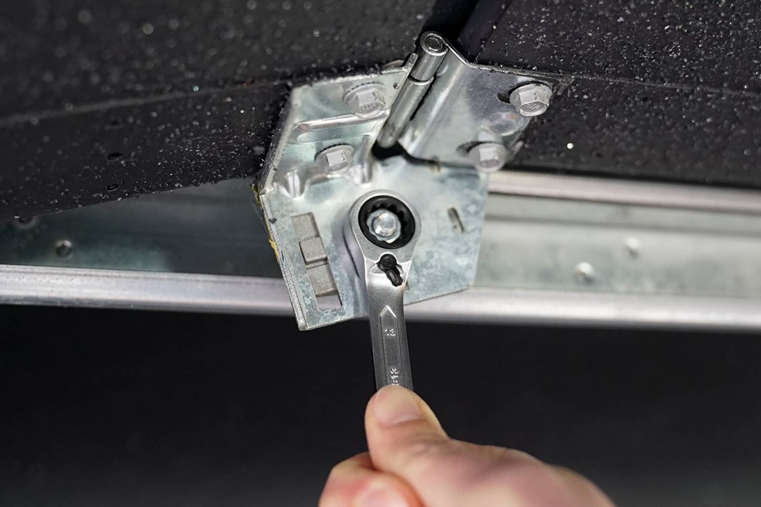 A hand is tightening a screw with a spanner on a metal bracket against a black surface.
