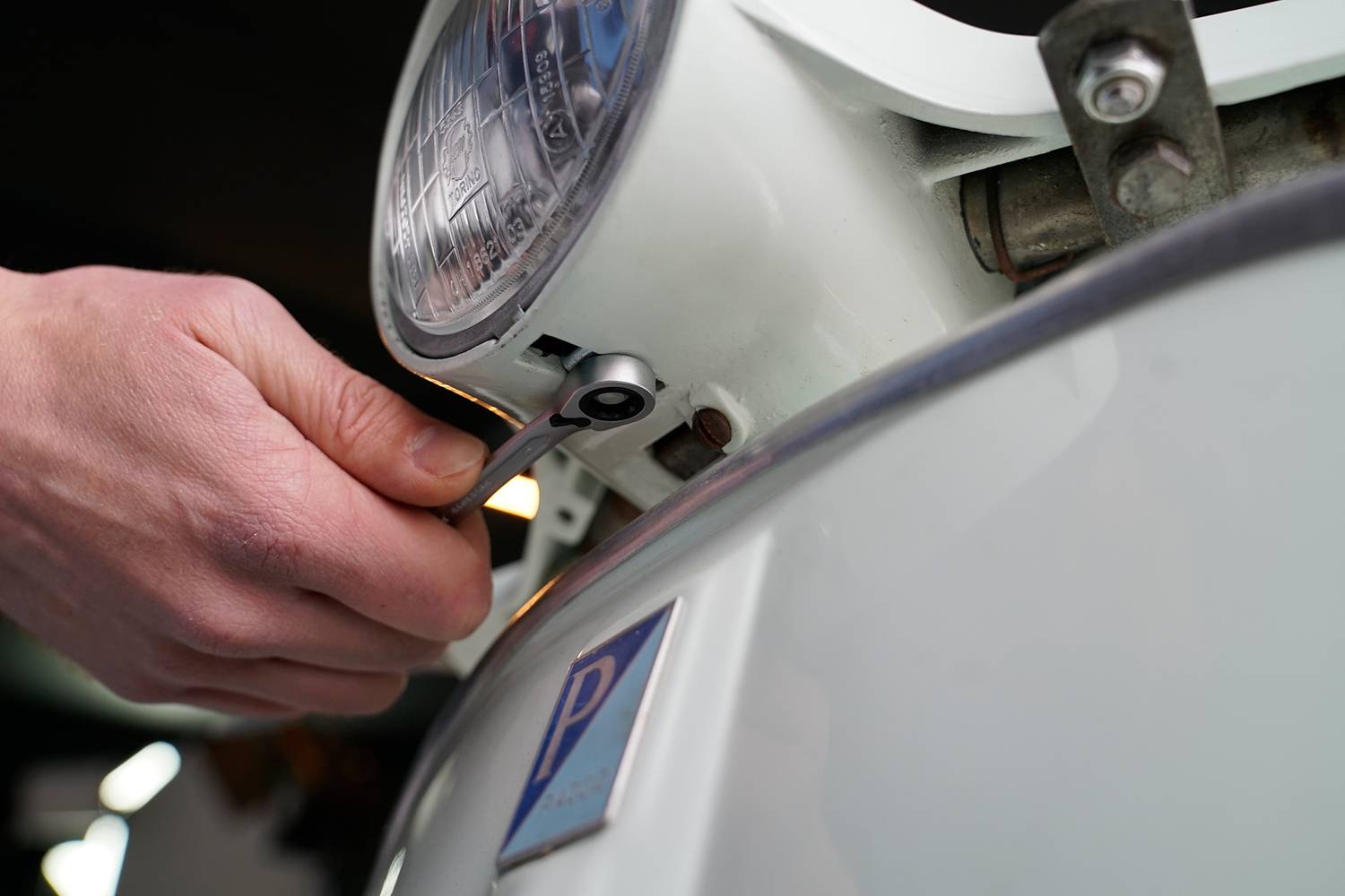A person is adjusting a component with a tool under the headlight of a white vehicle.