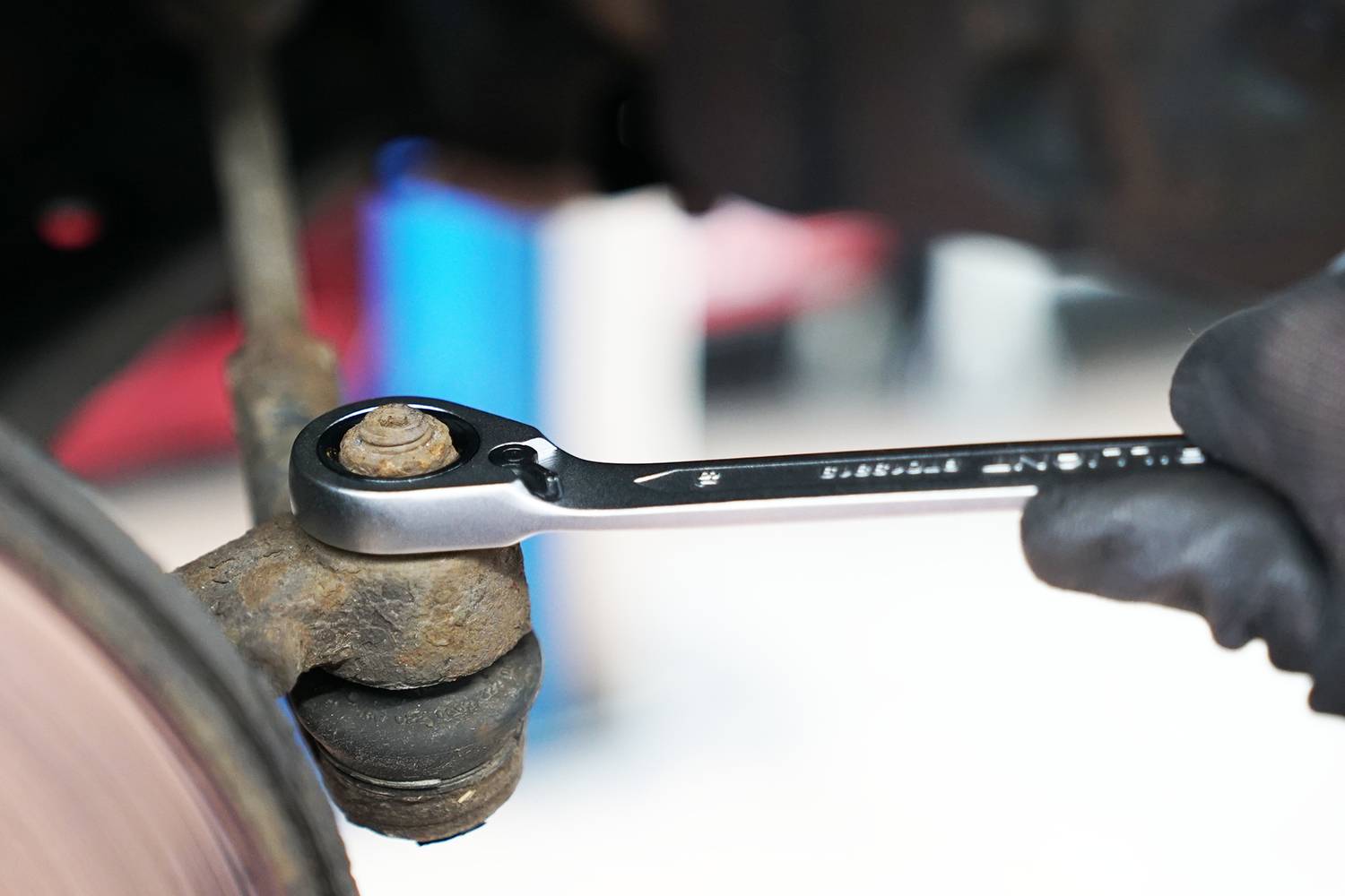 A hand is operating a spanner to turn a rusty bolt on a vehicle part. The car is being repaired.