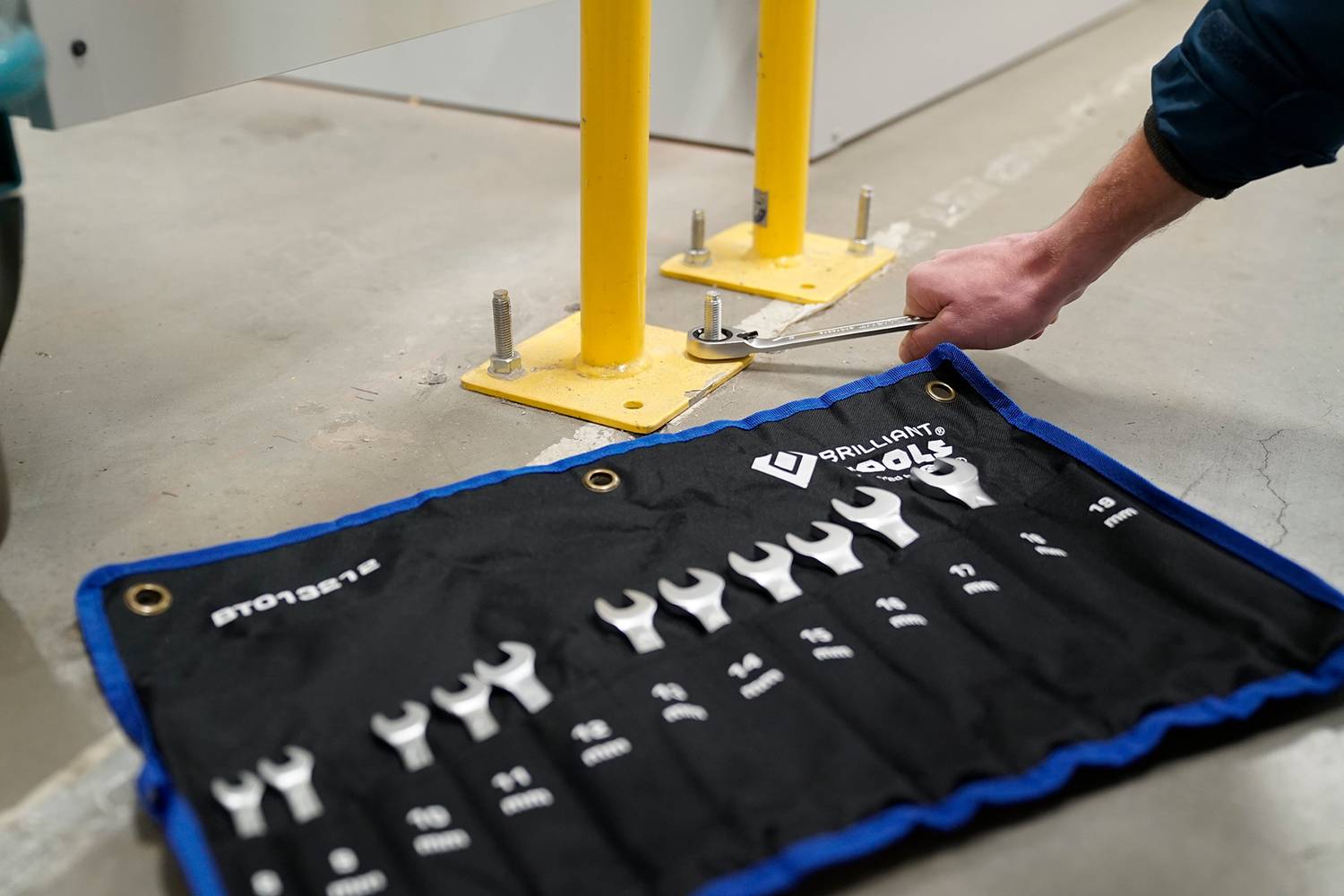 A technician tightens a yellow support rod with a spanner. Next to him lies an open toolkit with various spanners.