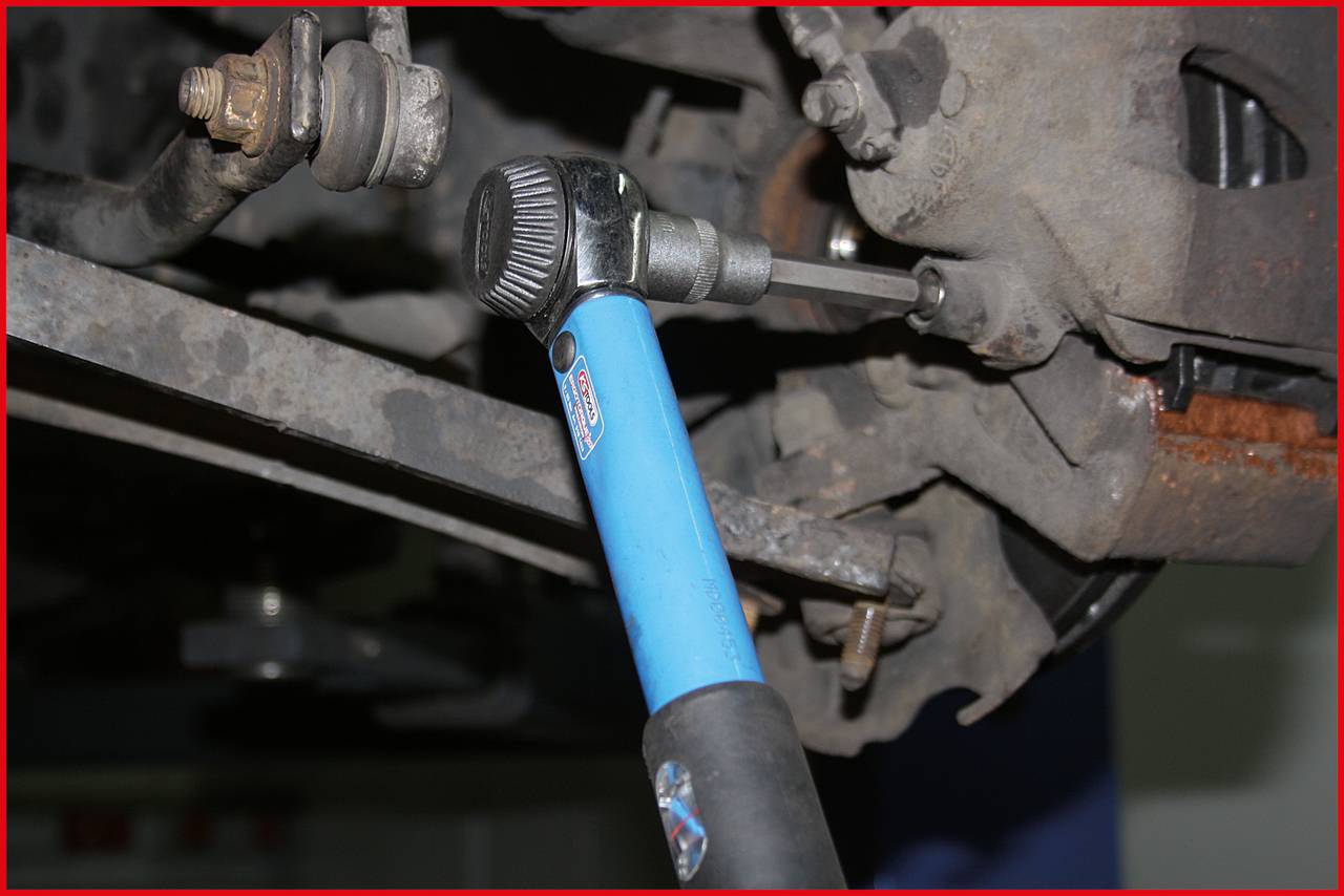 A torque wrench tightens a bolt on a car's rear axle. The suspension components are partially visible.