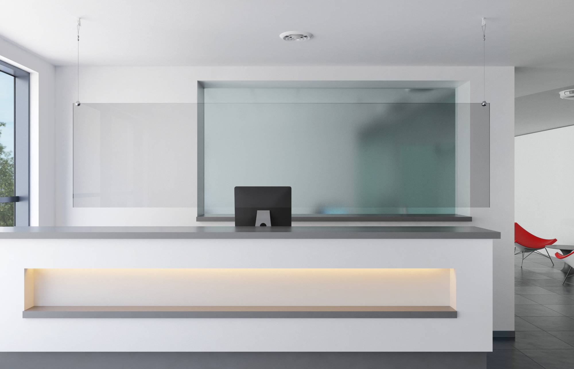 'A clean, modern reception desk with a computer monitor, glass partition, and natural light from large windows; red chair visible in the background.'
