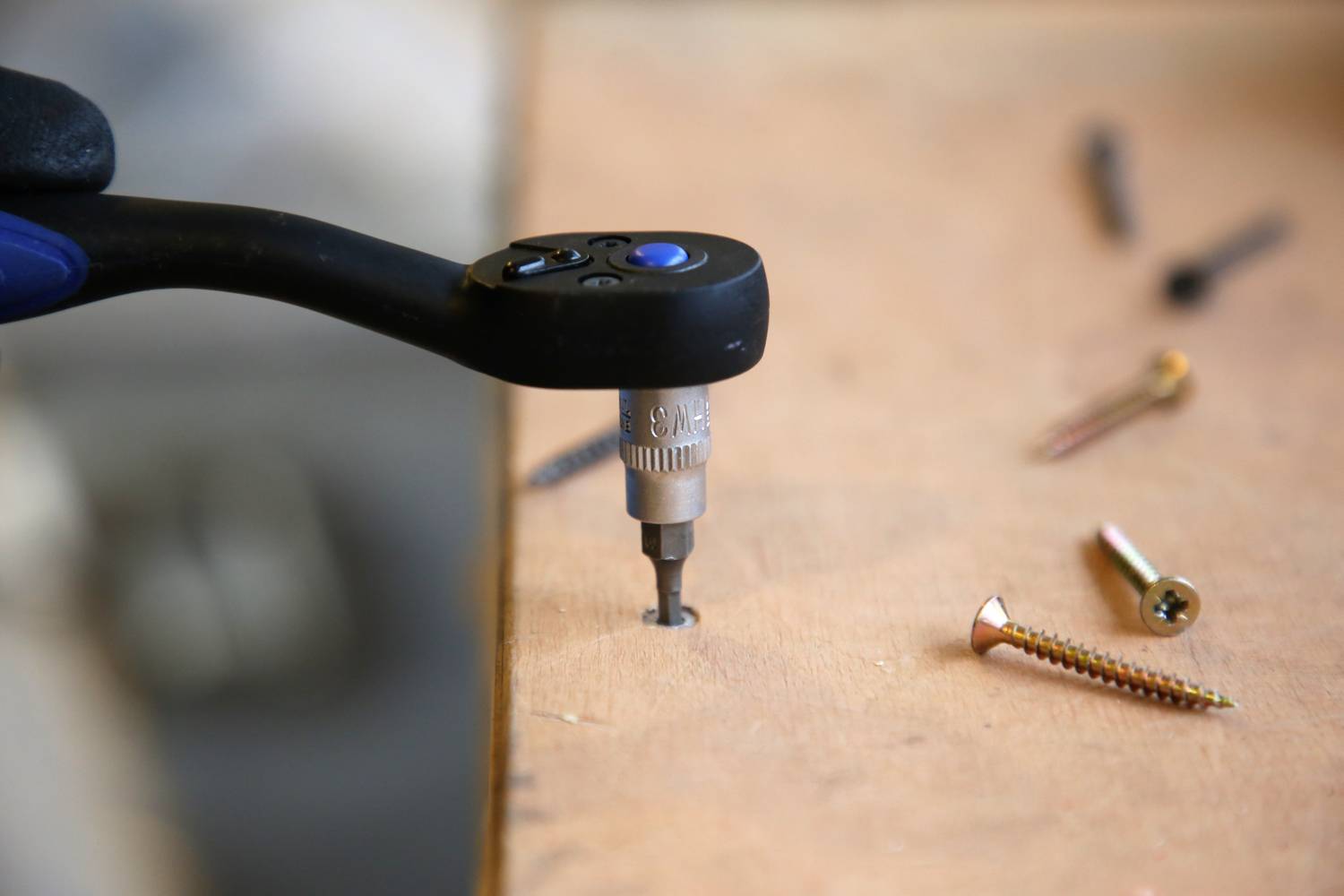 A cordless drill drives a screw into a wooden board. Several screws are scattered across the surface nearby.