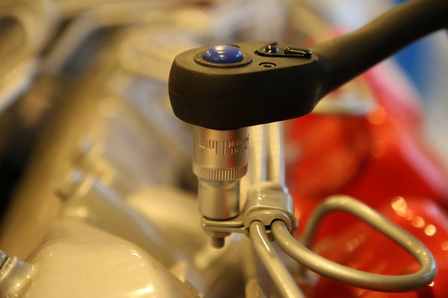 A ratchet tightens a screw on an engine. The tool has a blue button and is directed towards a metallic component.