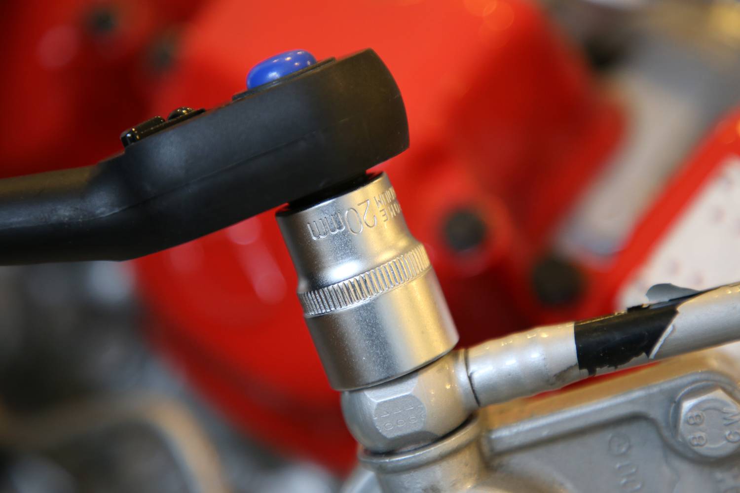 Close-up of a tool in use, tightening a metal nut on a machine. Background blurred with red components.
