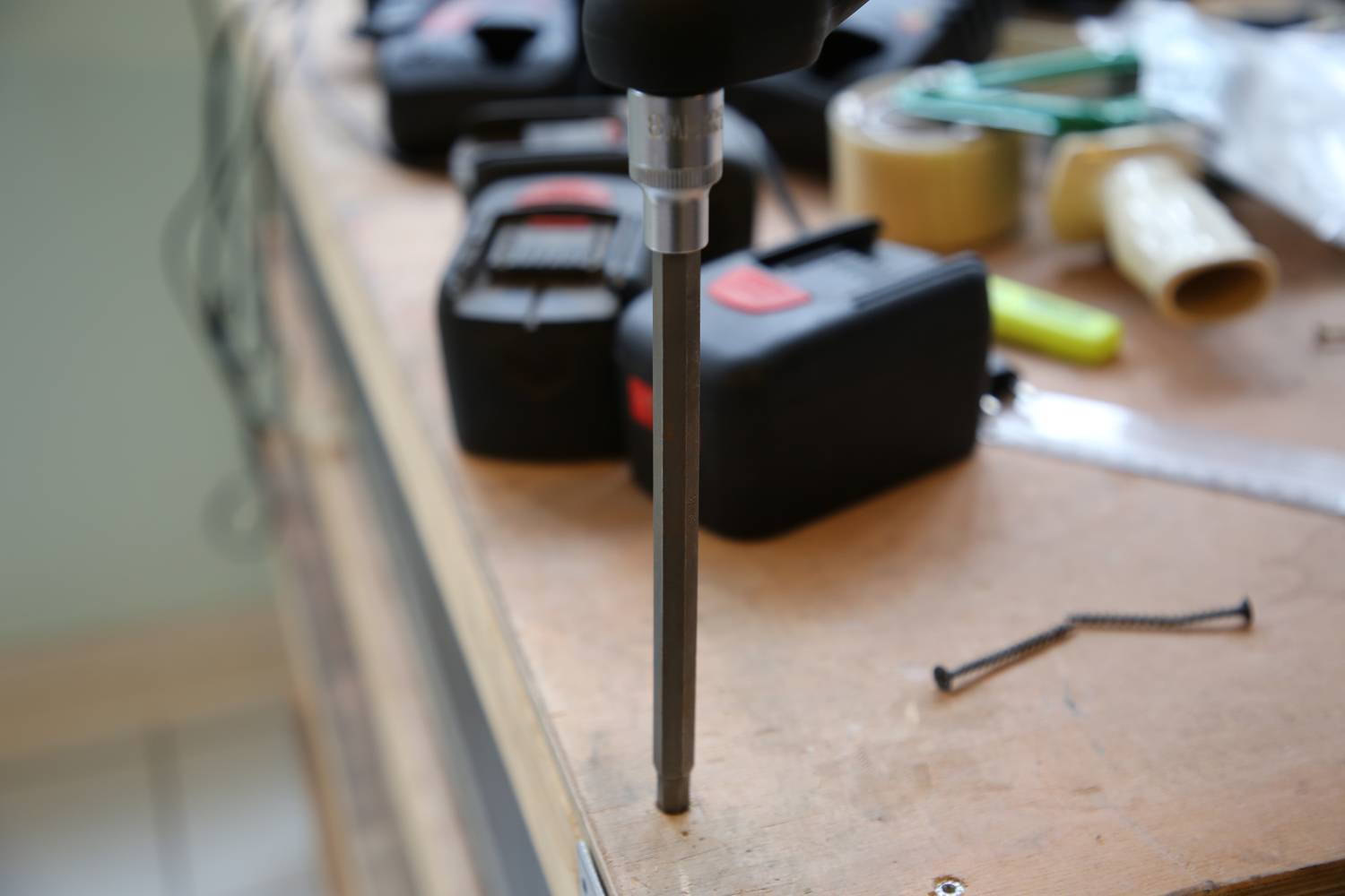 'A cordless drill creates a hole in a wooden surface on a workbench with screws and tools in the background.'