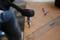 A person is drilling a hole in a wooden board using an electric drill. A transparent ruler with markings lies beside it.
