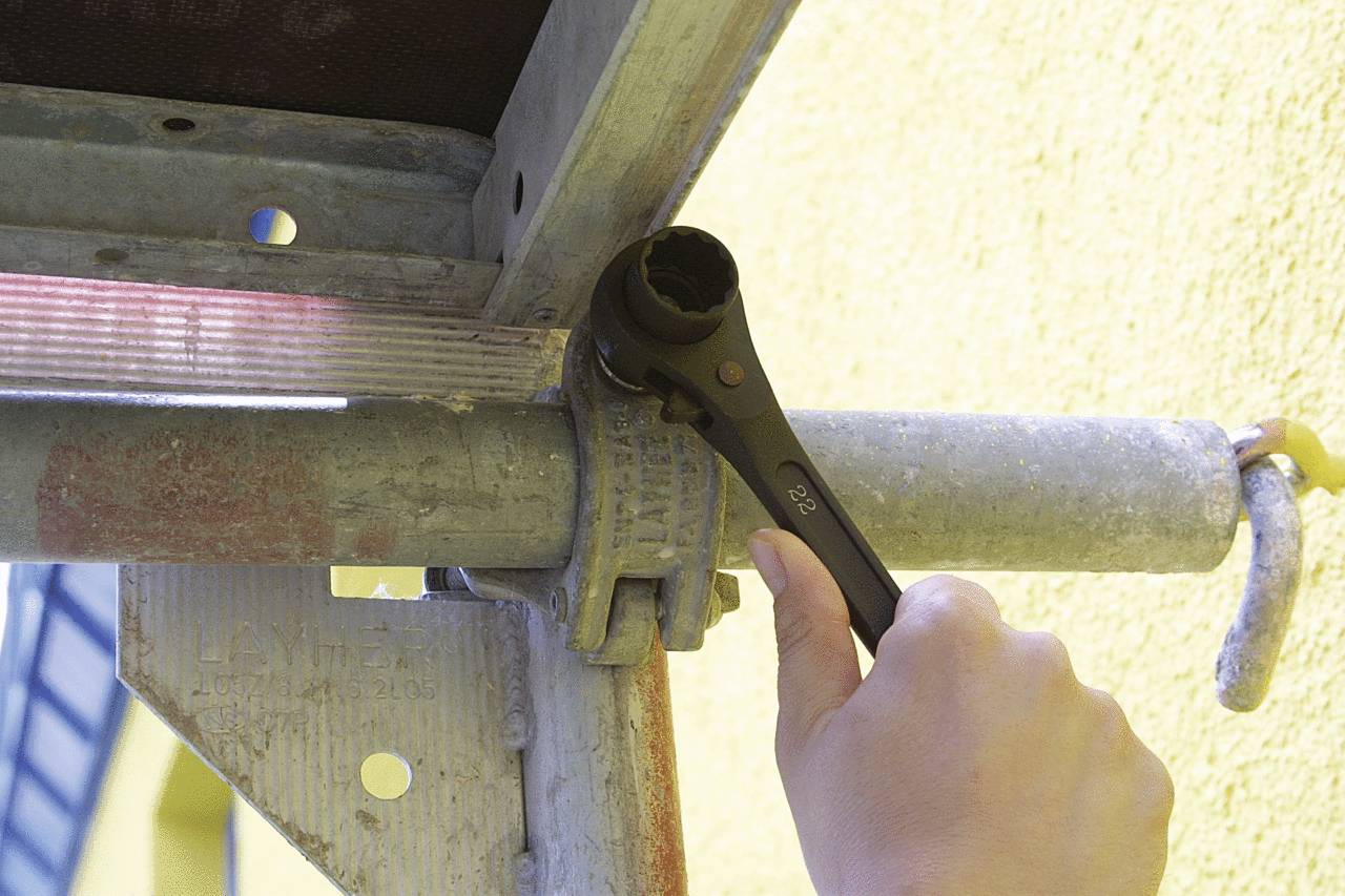 A hand tightens a nut with a spanner on a scaffolding, with a yellow wall in the background.