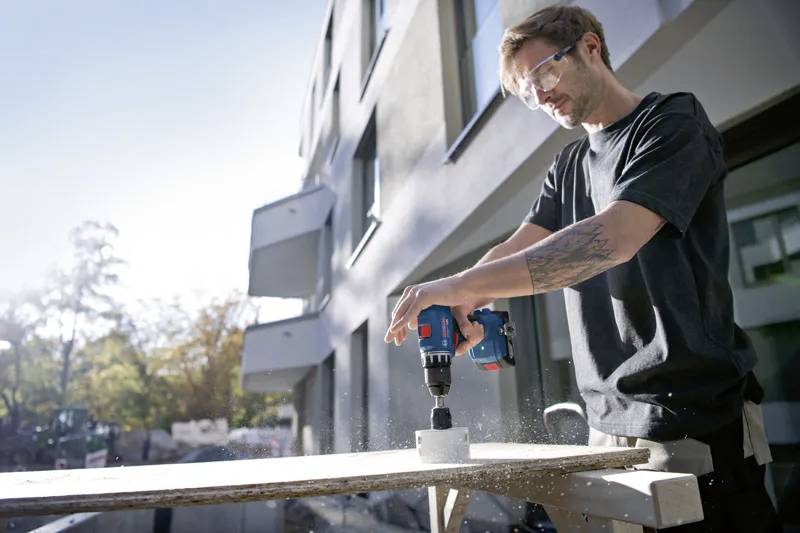 A person uses a power drill to cut a hole in a wooden board on a sunny day, wearing safety glasses, with buildings in the background.