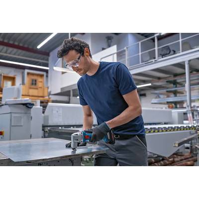 Worker wearing safety glasses and gloves cuts a metal sheet with a handheld power tool in an industrial workshop.