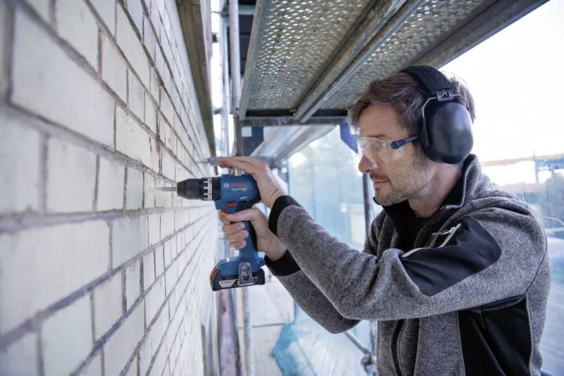A person wearing protective gear uses a drill on a brick wall, with scaffolding and construction materials in the background.