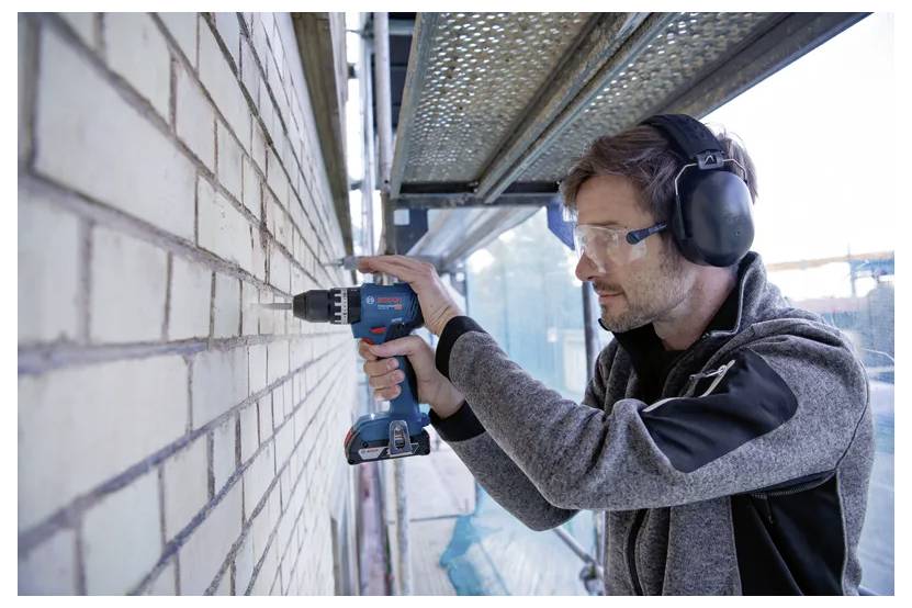 A man wearing safety goggles and earmuffs uses a power drill on a brick wall at a construction site, highlighting safety in construction work.