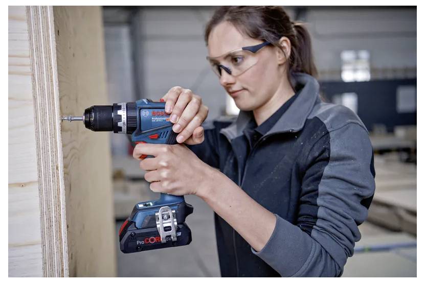 A woman wearing safety glasses uses a cordless drill to fasten screws into wooden boards in a workshop setting.