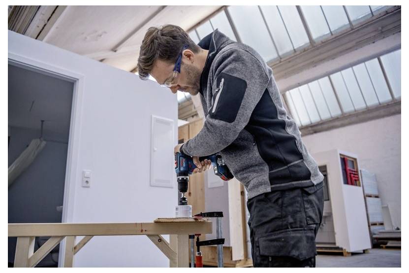 A person using a power drill to cut a circular hole in a wooden board, inside a well-lit workshop with industrial decor.