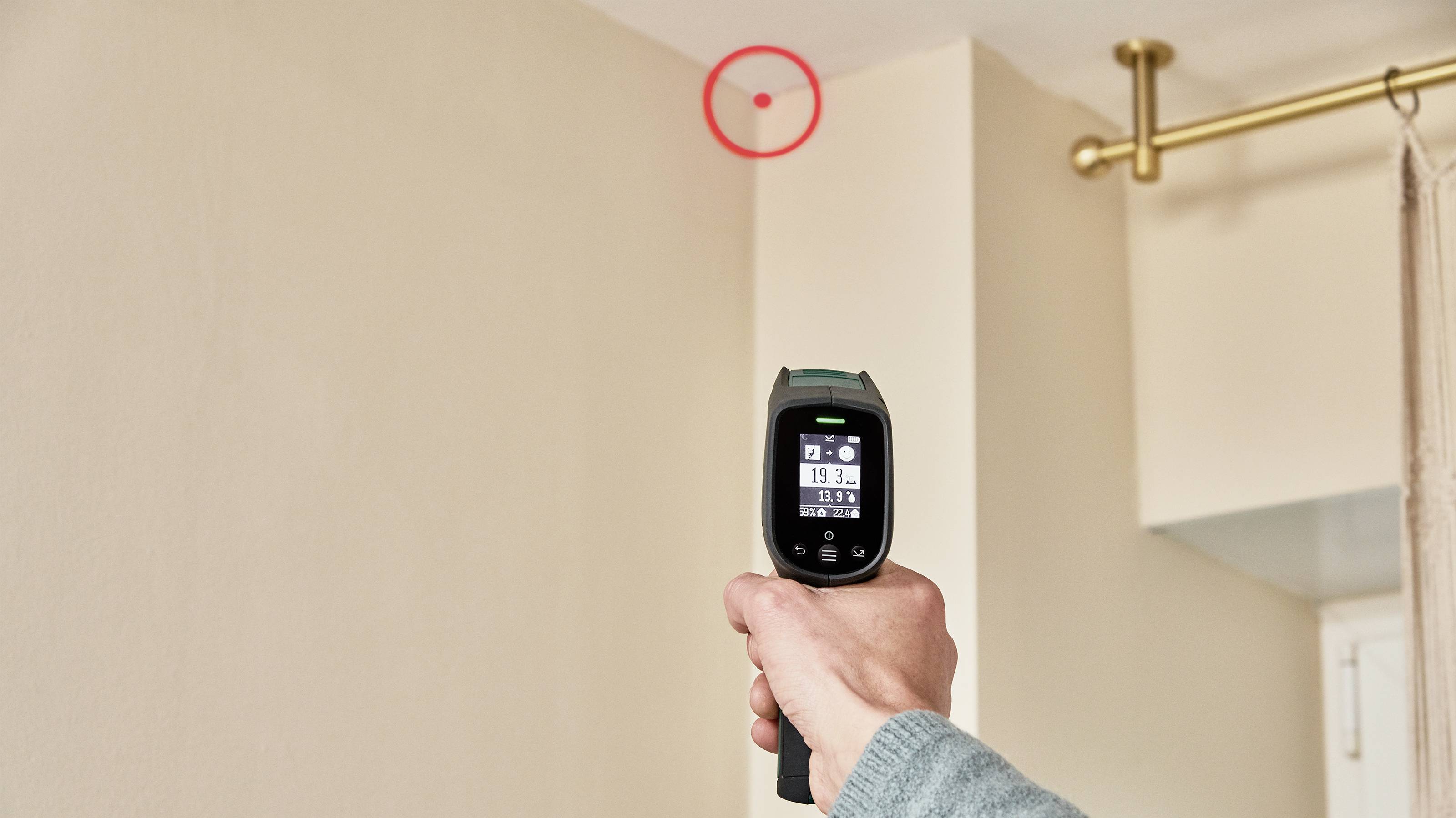 A person aims an infrared thermometer at a red mark on the wall to measure the temperature.