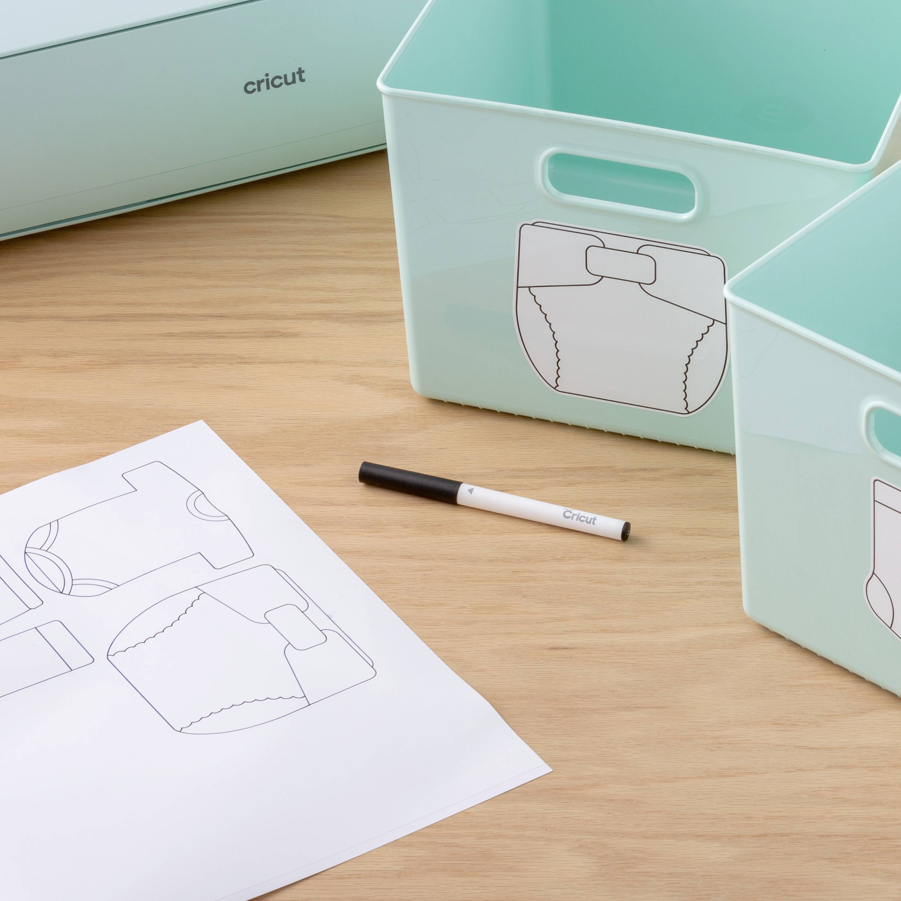 Two mint-coloured boxes with a nappy motif printed on a wooden surface. To the right, a drawing pencil lies next to a sketch of nappies.