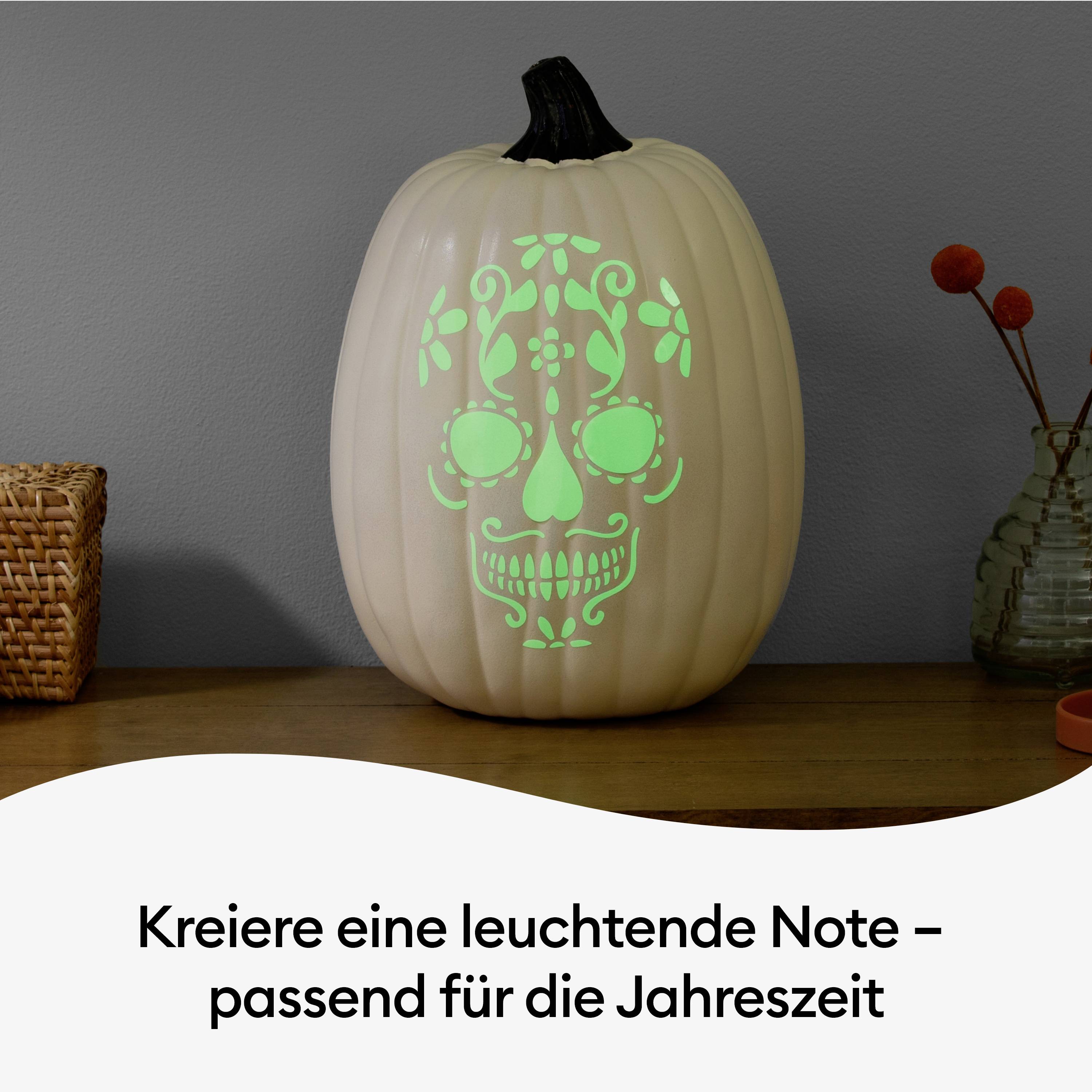 A white pumpkin displays a glowing, decorative facial pattern. Beside it, a basket and a vase sit on a table.