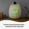 A white pumpkin displays a glowing, decorative facial pattern. Beside it, a basket and a vase sit on a table.