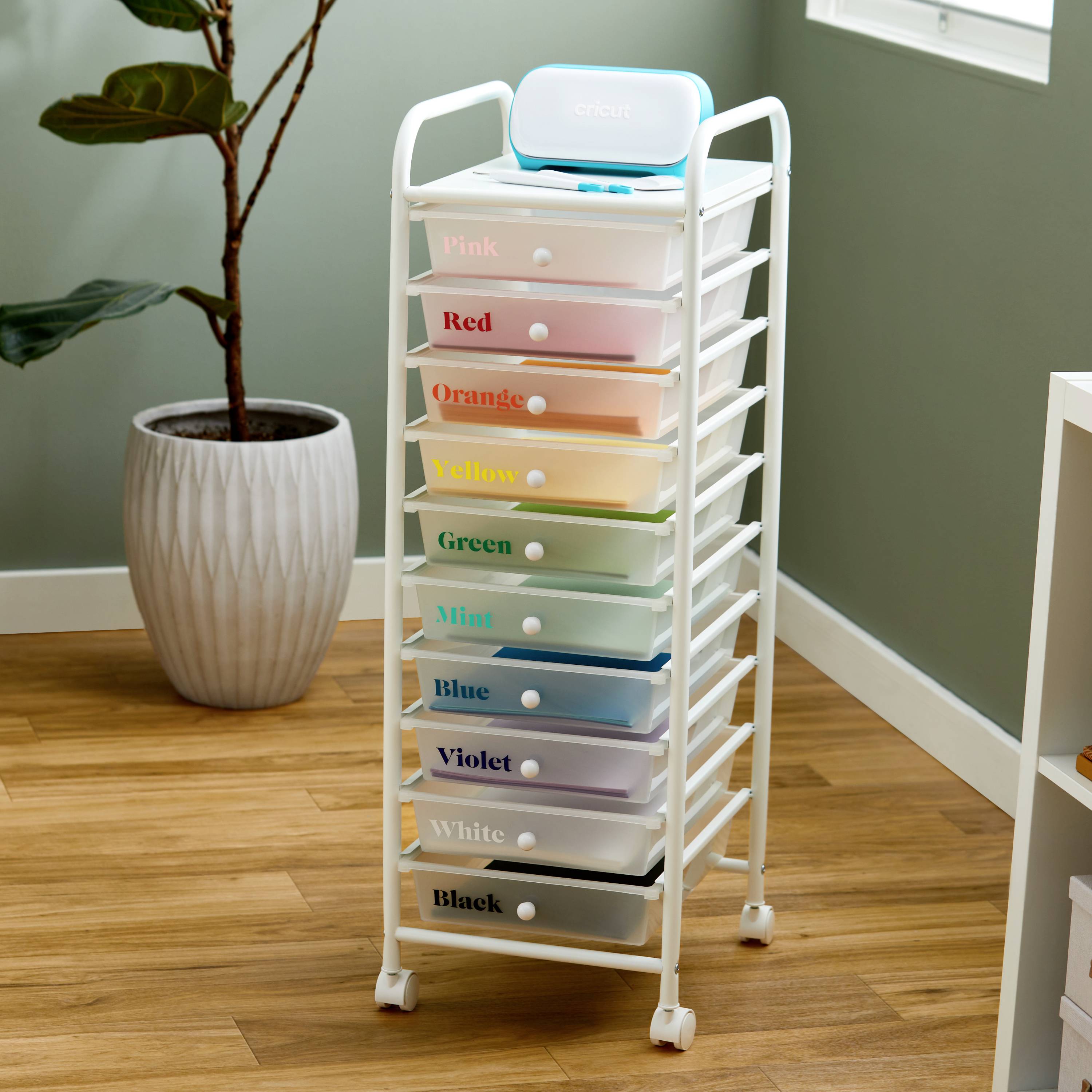A white roller container with nine coloured drawers, each labelled from 'Pink' to 'White', stands next to a plant on a wooden floor.