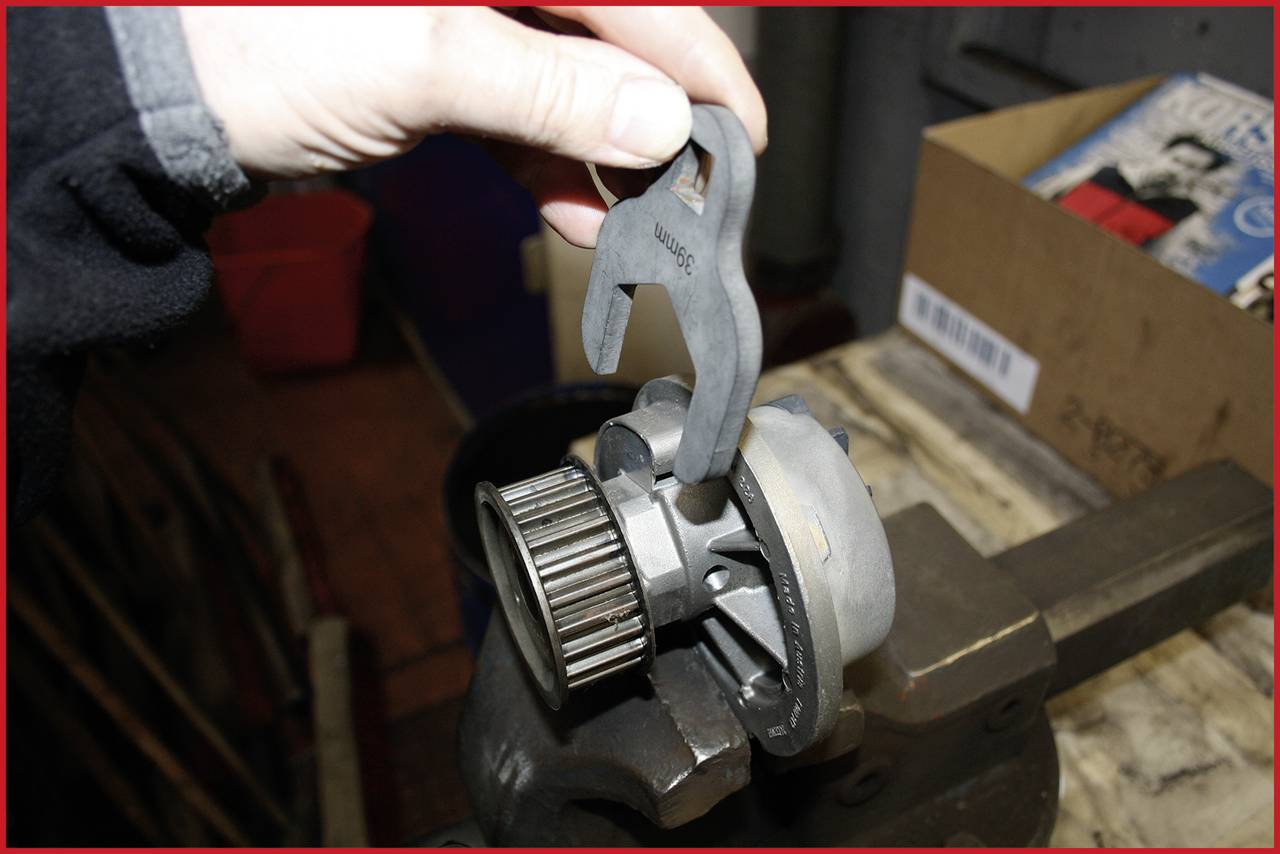 A hand holds an adjustable wrench to adjust a car part holder in a vice. A workshop is in the background.